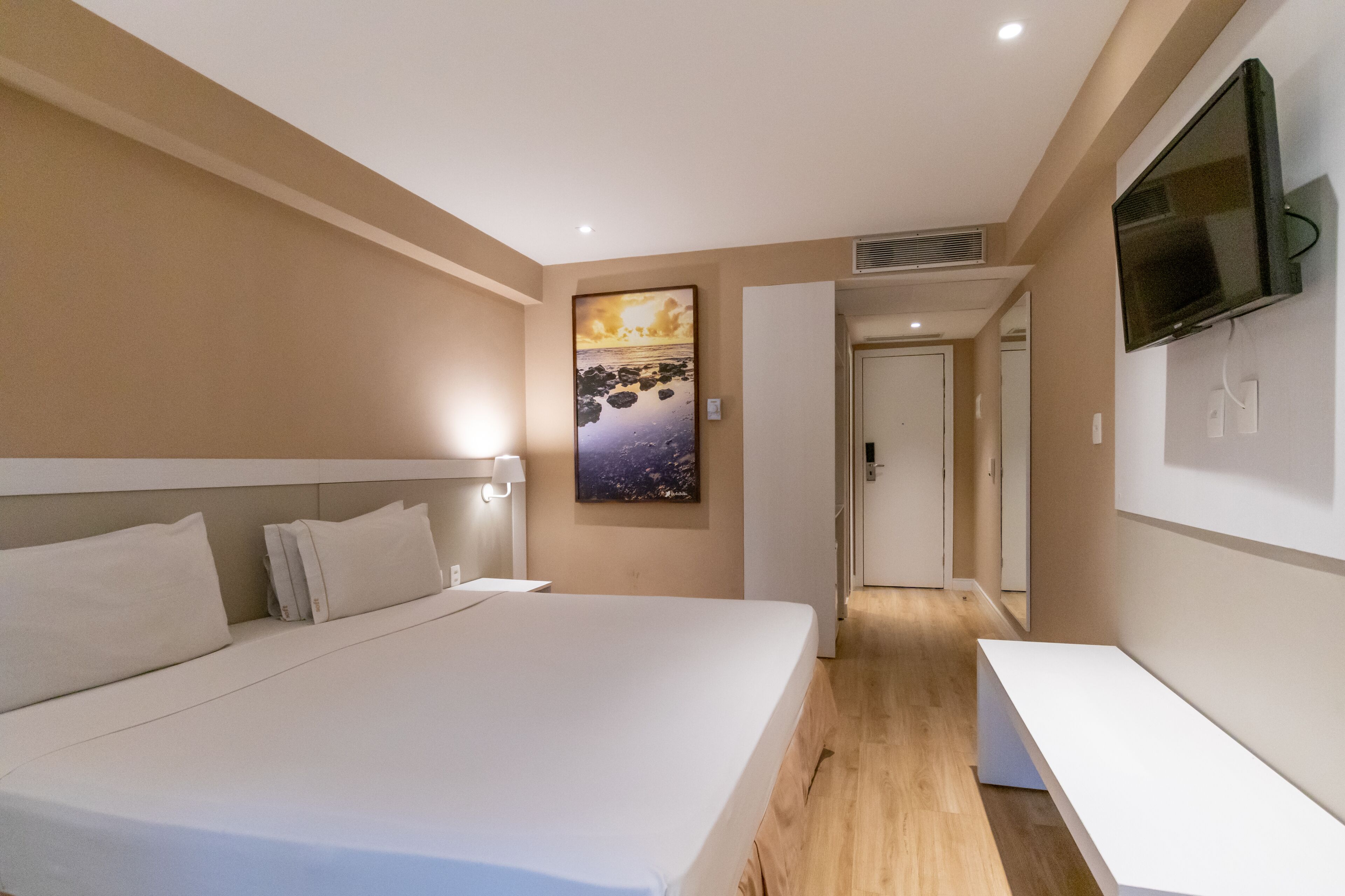 Quarto de hotel moderno com uma cama grande, televisão de tela plana na parede e um quadro sobre a cama.