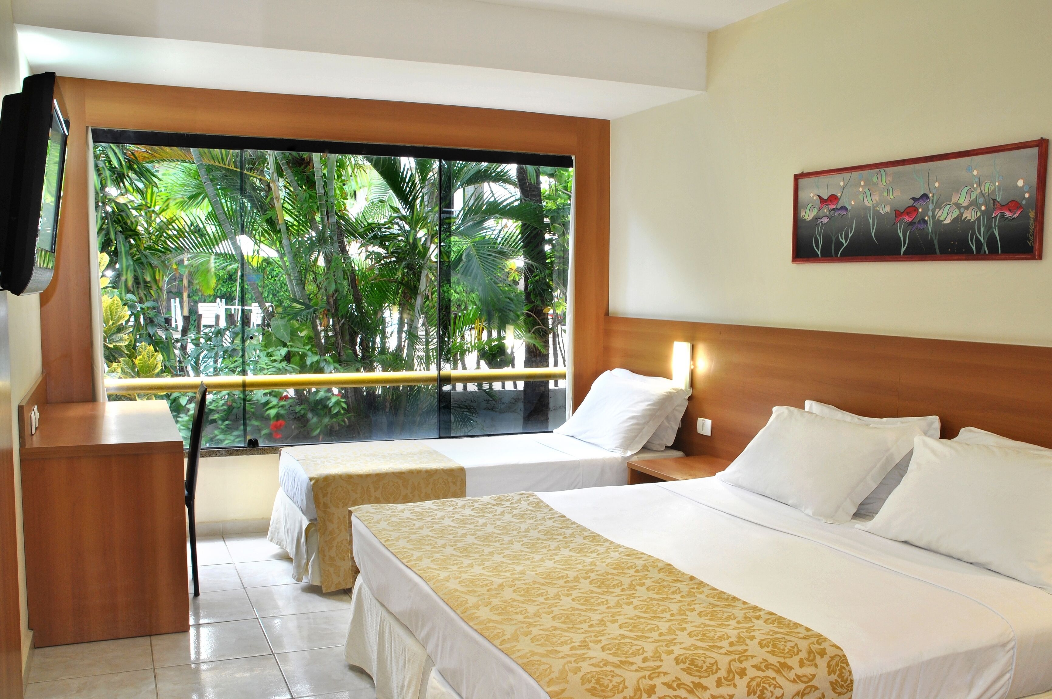 A hotel room with two beds, a desk, and a large window with a view of tropical foliage.