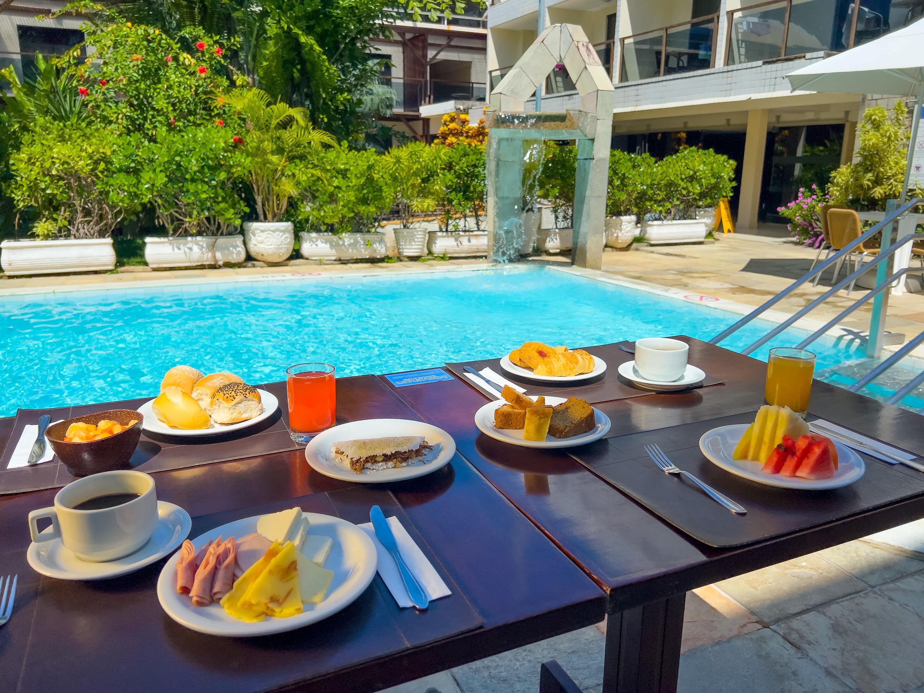 Une table à manger, dressée avec des aliments pour le petit-déjeuner, se trouve au bord de la piscine. On y voit des assiettes garnies de viennoiseries, de fruits et de boissons.