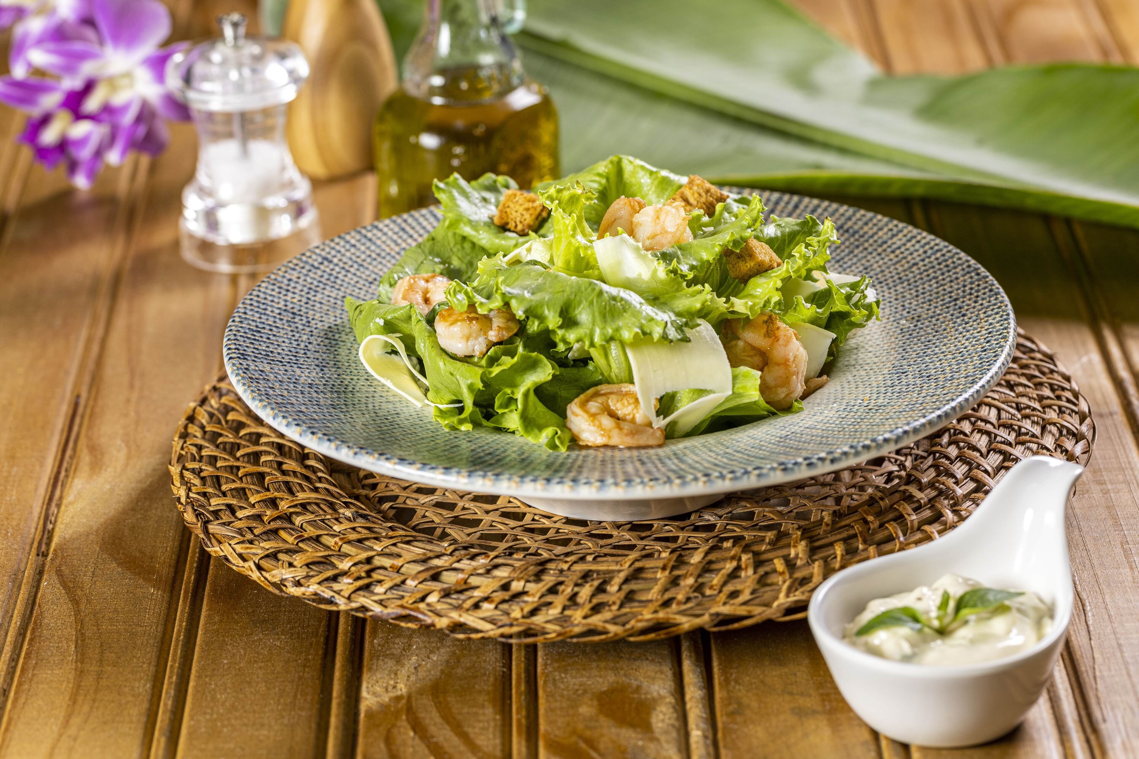 A plate of shrimp salad on a woven placemat, with a small bowl of dressing and a salt shaker in the background.