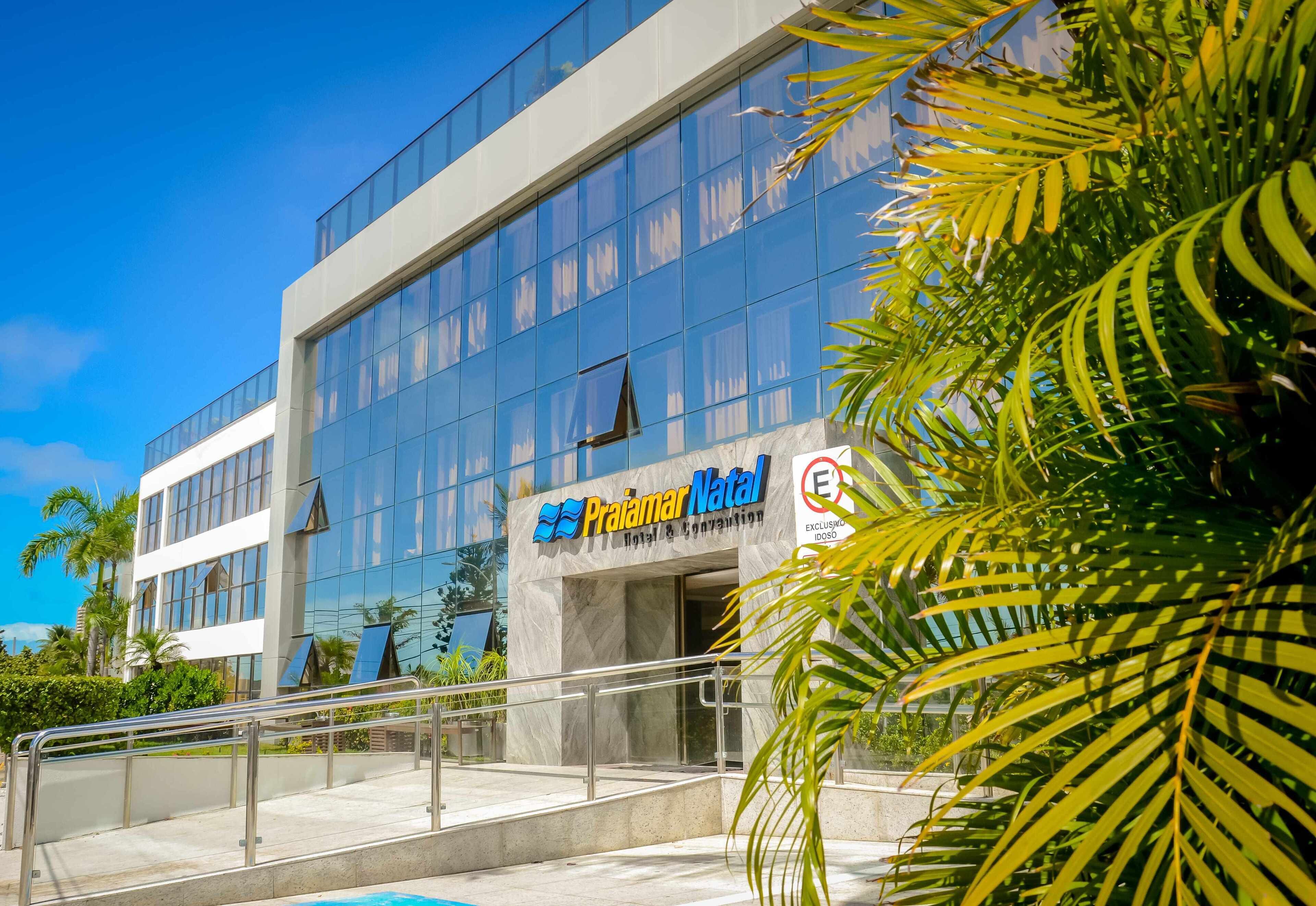A modern building with a glass facade, a sign for 'Praiamar Natal Hotel & Convention', and a palm tree in the foreground.