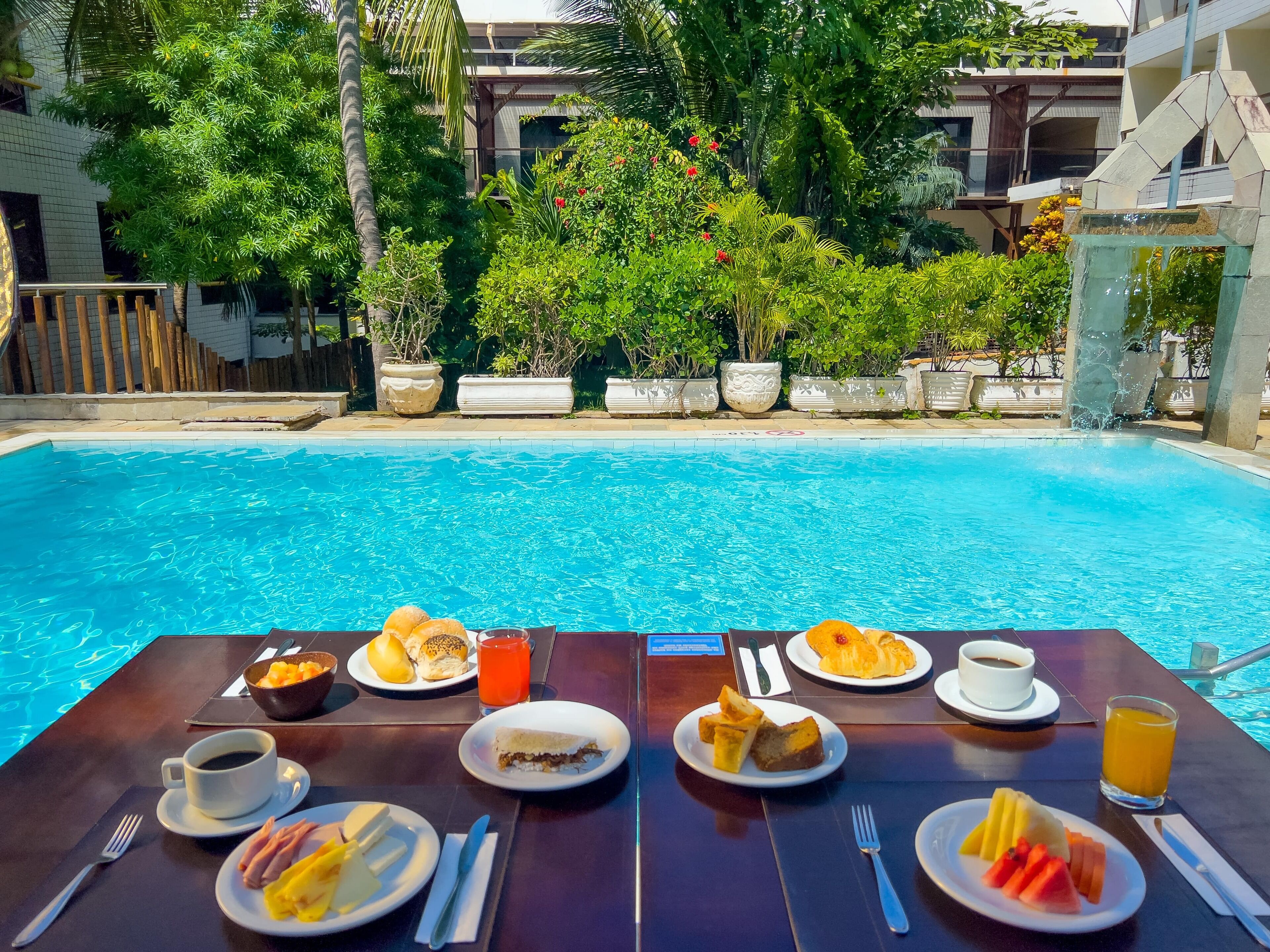 Mesa de refeições à beira da piscina, com itens de café da manhã e vista para uma piscina cercada por vegetação exuberante e um edifício.