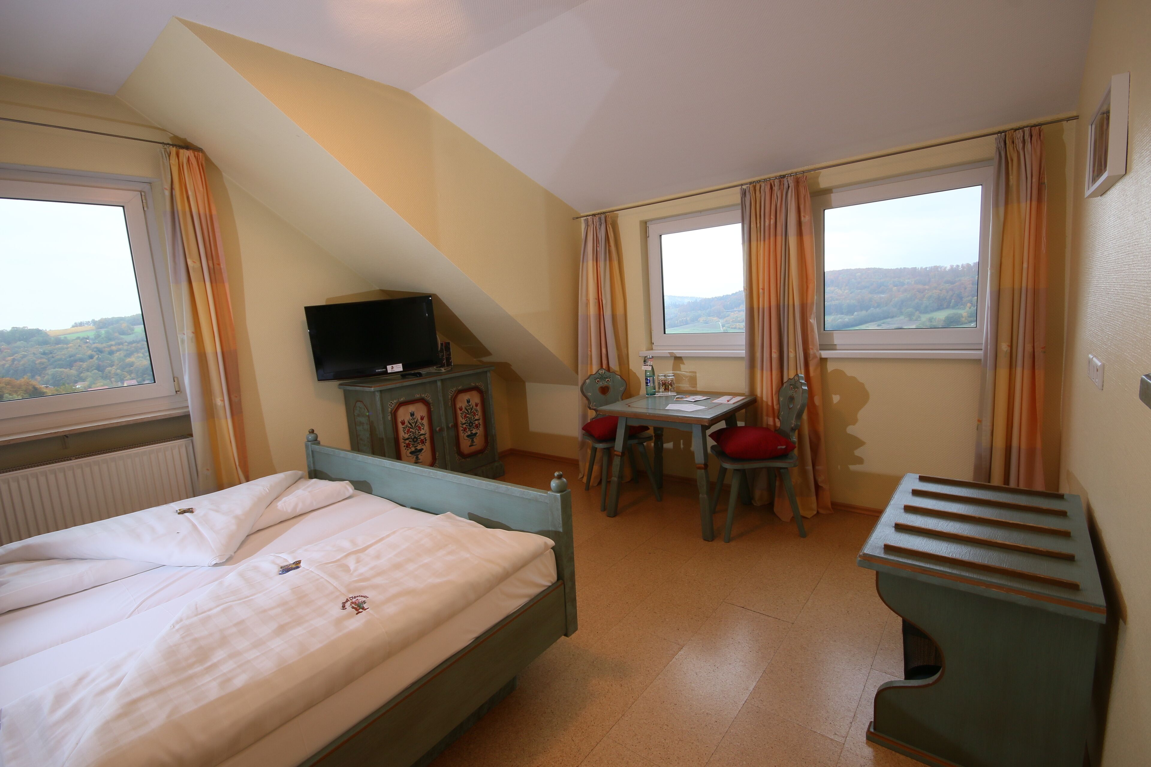 A bedroom with a bed, a desk, a chair, a television, and a window with curtains.