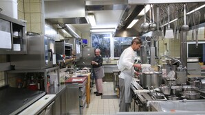 Una cocina profesional con electrodomésticos de acero inoxidable, utensilios colgantes y dos chefs trabajando.