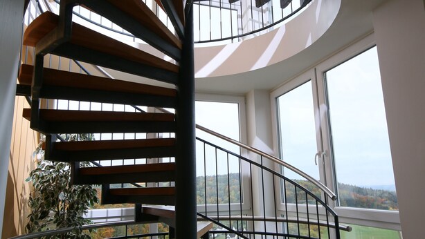 Una escalera de caracol con escalones de madera y barandillas negras, que conduce a un balcón con vistas al campo.