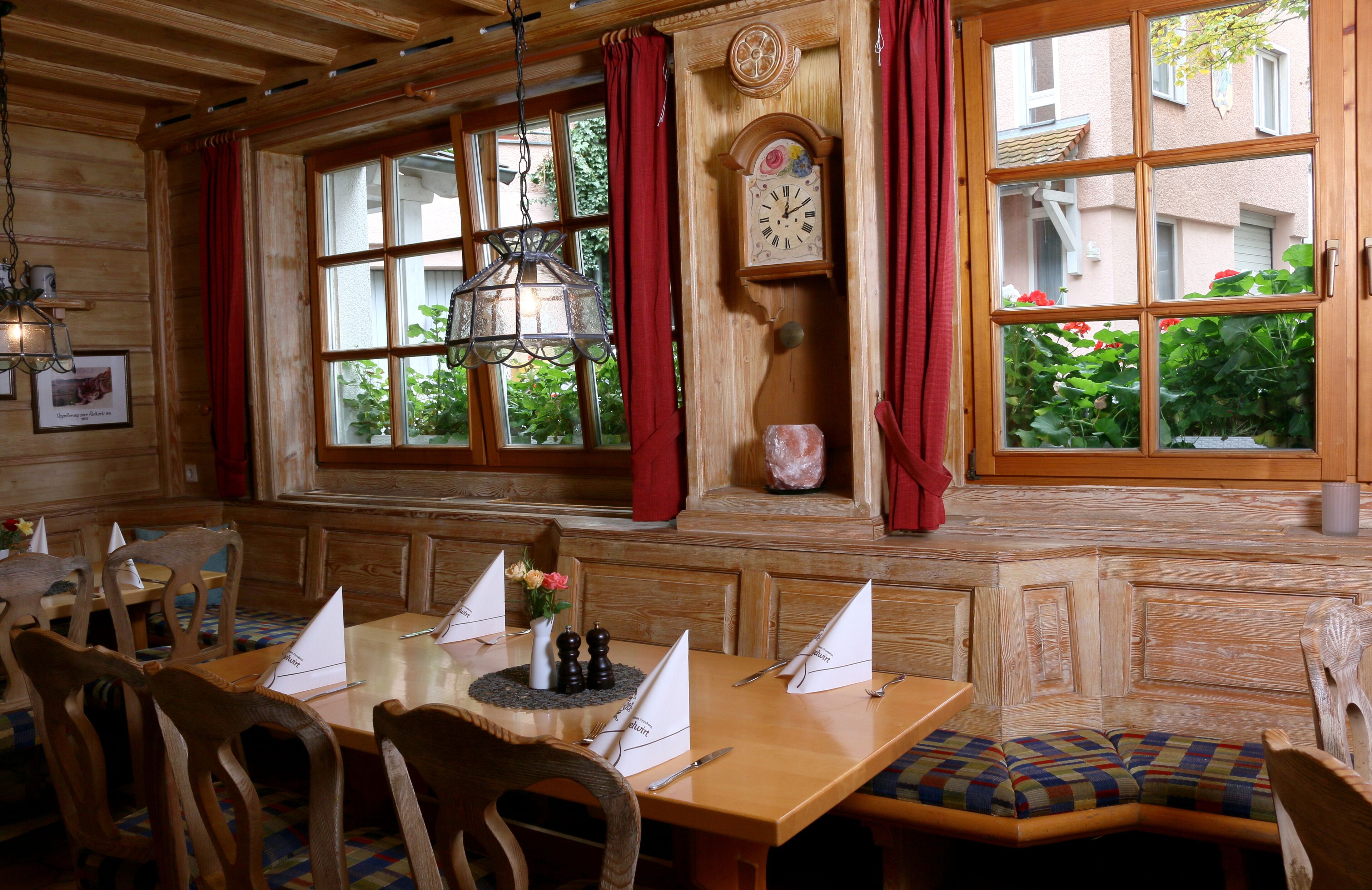 Un interior de madera con una mesa de comedor preparada para dos, un reloj y una ventana con cortinas rojas.