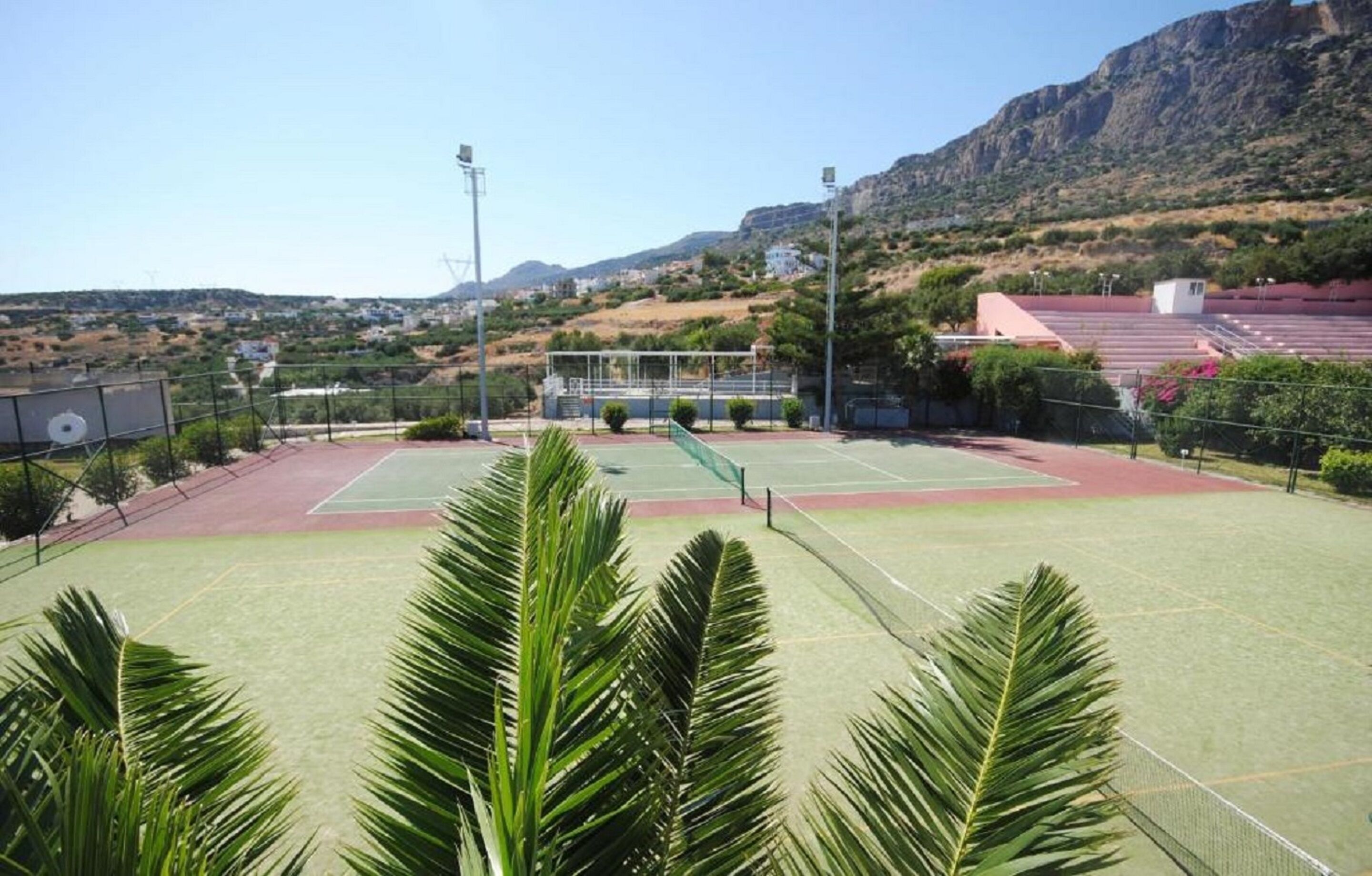 Una cancha de tenis al aire libre con red, rodeada de palmeras y montañas al fondo.