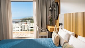 Une chambre d’hôtel avec un lit, un bureau et une chaise, une petite table ornée d’un vase de fleurs, et une vue sur la mer depuis un balcon.