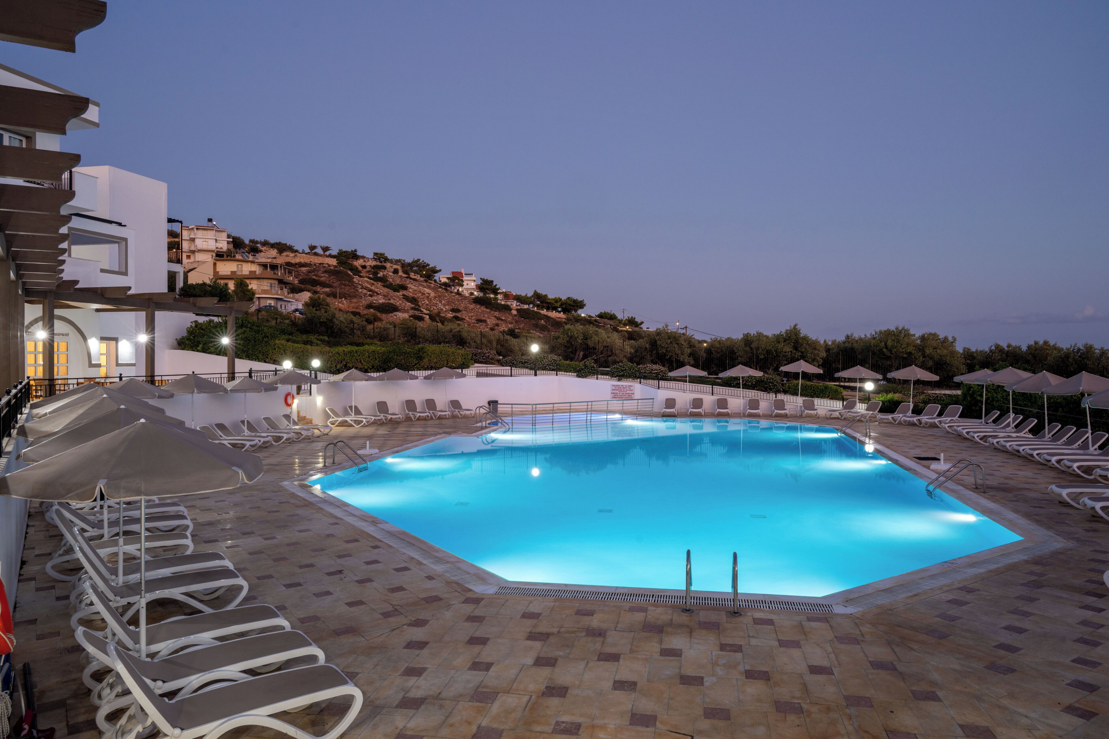 A swimming pool with lounge chairs and umbrellas, surrounded by a tiled deck and buildings in the background.
