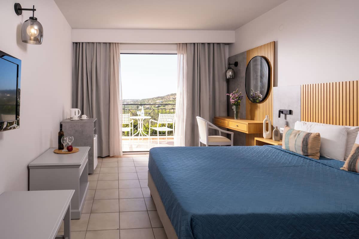 Une chambre d’hôtel avec un lit, un bureau, une chaise, une petite table avec une bouteille de vin et un verre, et une vue sur l’extérieur.
