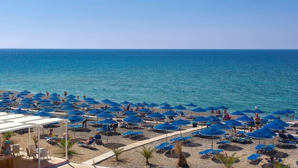 Une plage bordée de nombreux parasols bleus et de chaises longues, une mer d’un bleu limpide et une rive sablonneuse.
