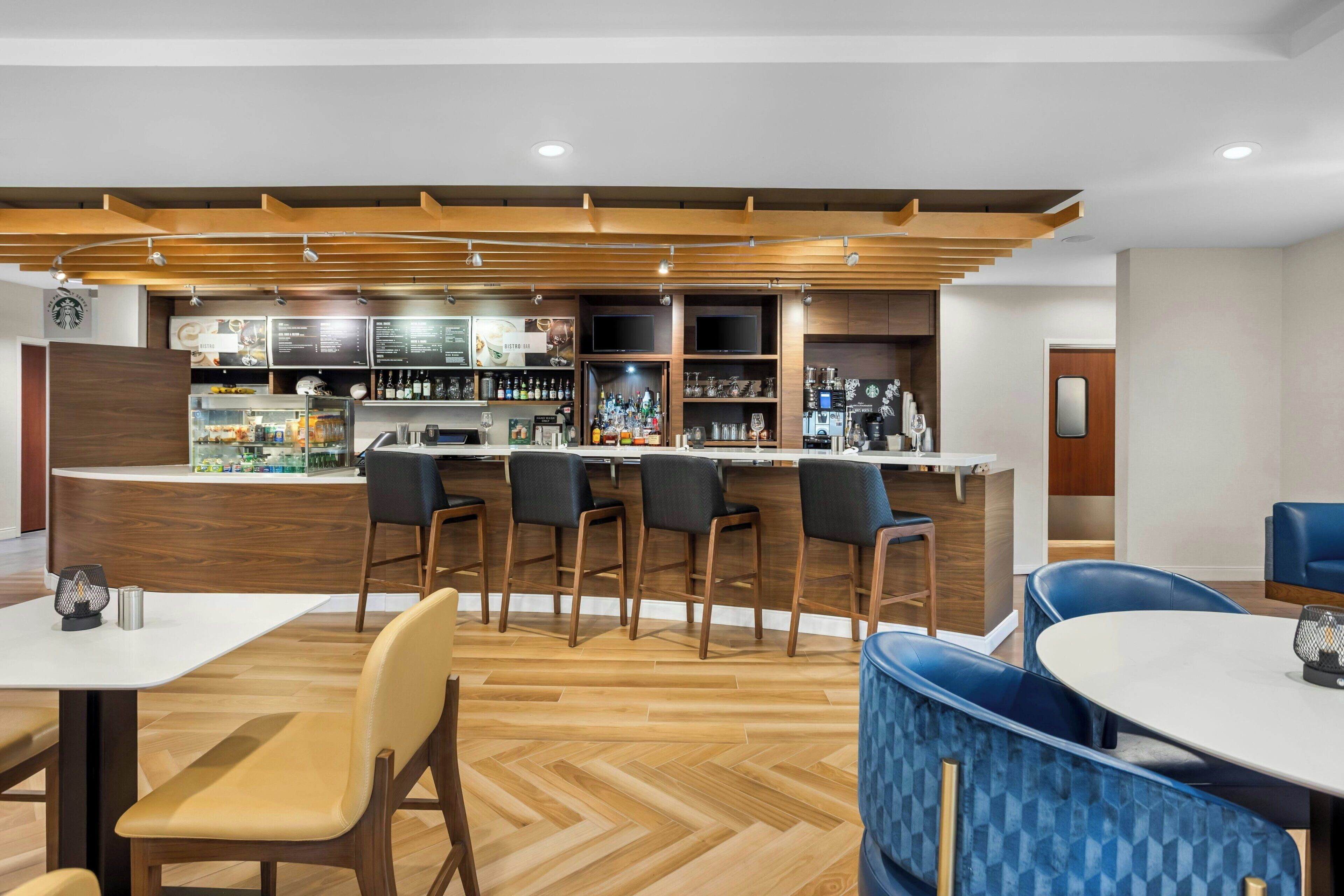 Zona de bar moderna con mostradores de madera, taburetes negros y una variedad de botellas y vasos.