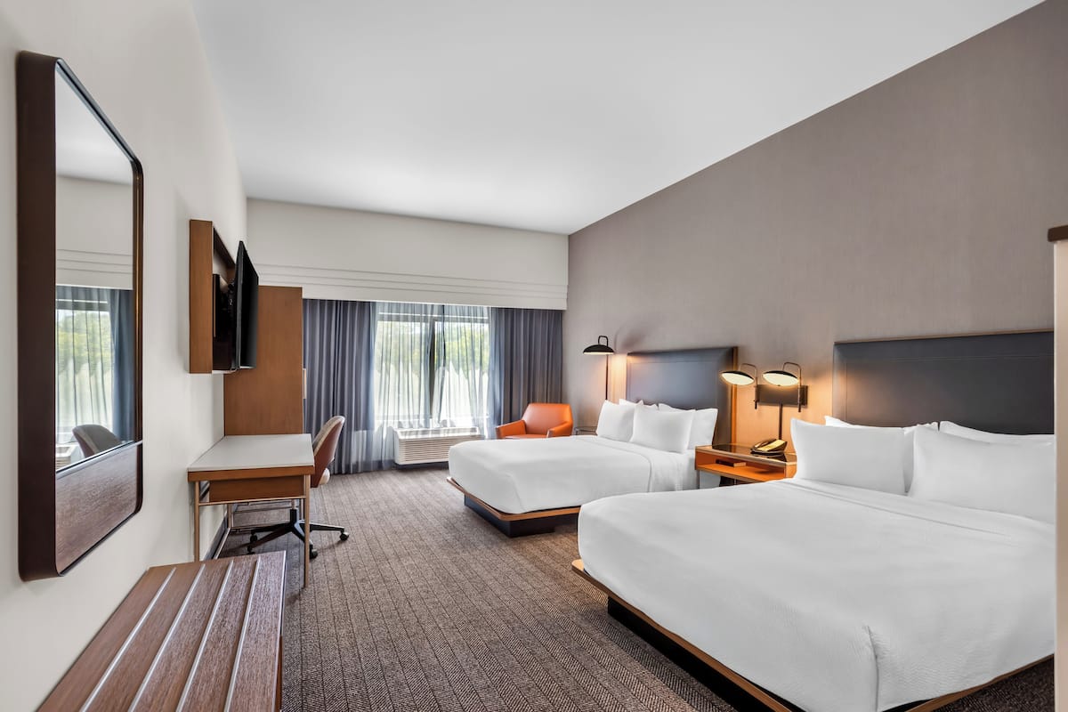 Habitación de hotel con dos camas, un escritorio, una silla, un televisor y un espejo grande.