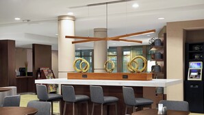 Un moderno vestíbulo de hotel con una zona de bar central, diversas opciones de asientos y un quiosco de autoservicio.