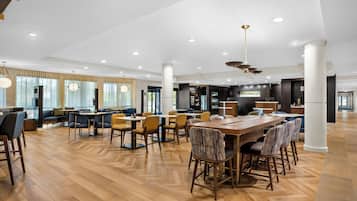 Un amplio vestíbulo de hotel con zona de comedor central, arreglos de asientos y un bar.