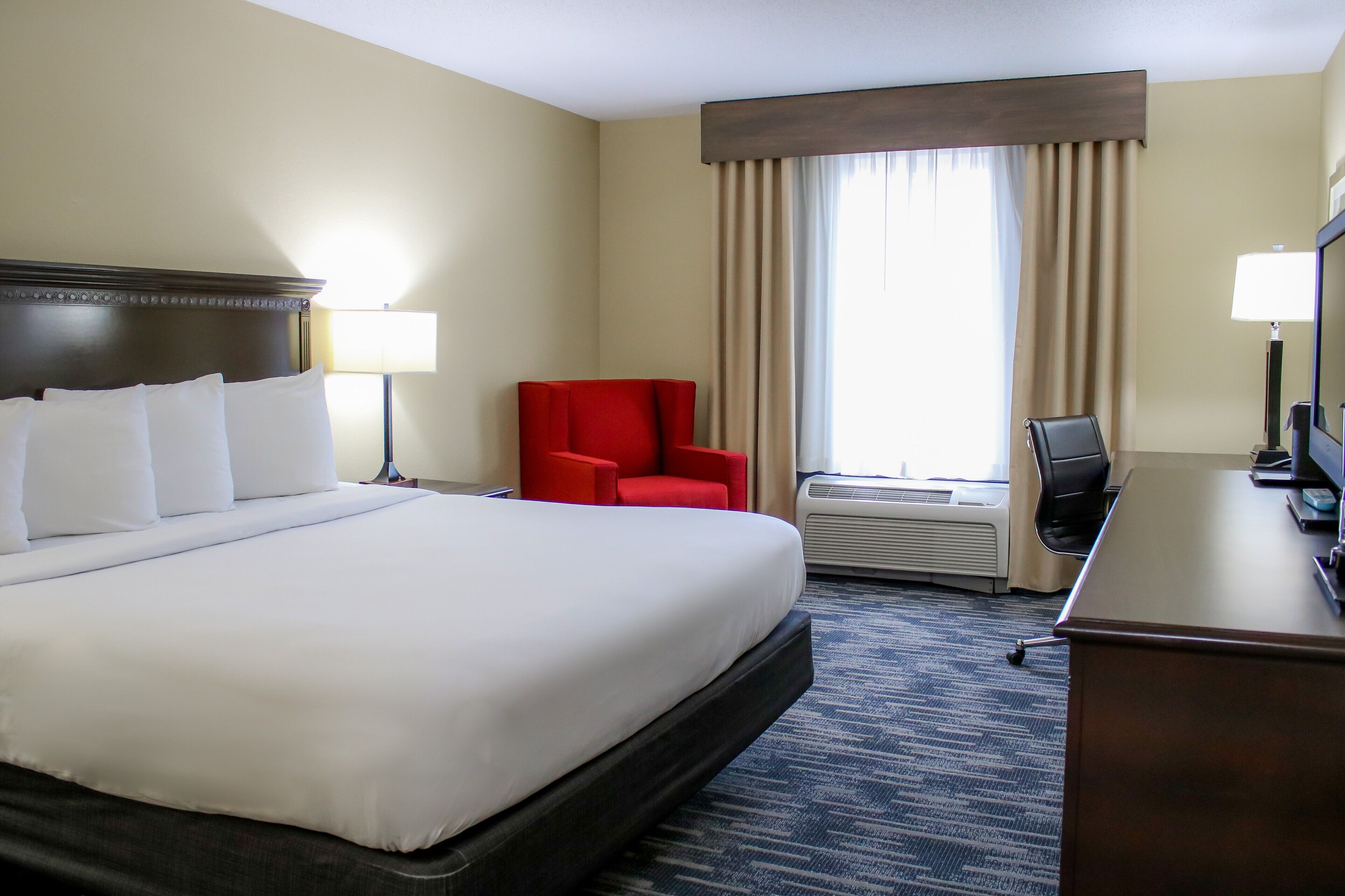 A hotel room with a large bed, a red chair, a desk with a computer, and an air conditioning unit.