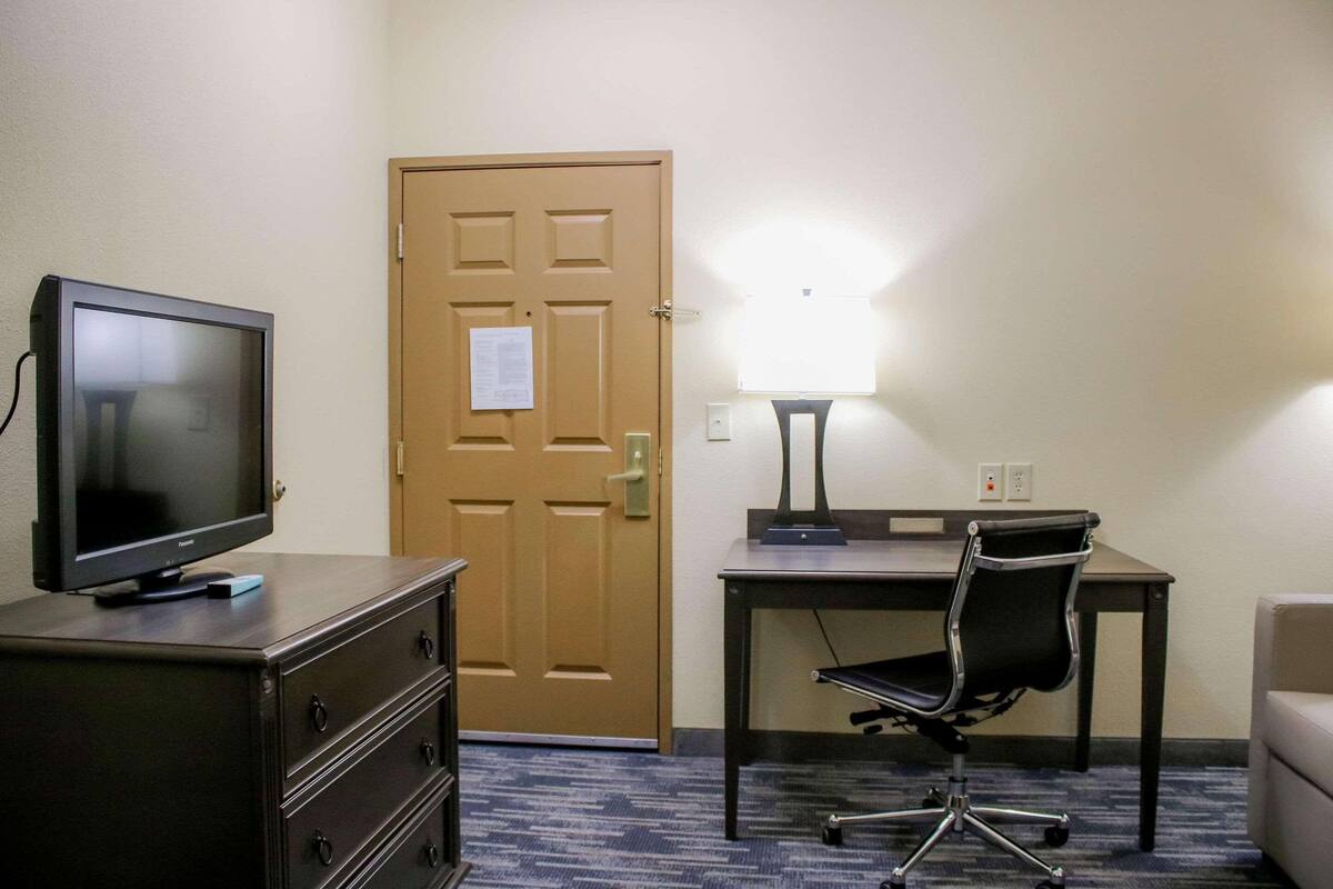 A hotel room with a television on a dresser, a desk with a chair, a lamp, and a door with a notice.