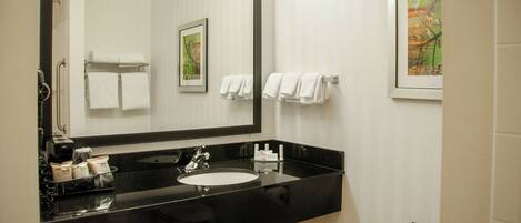 A bathroom with a black countertop, white sink, and a large mirror. There are towels hanging on a rack and a framed picture on the wall.