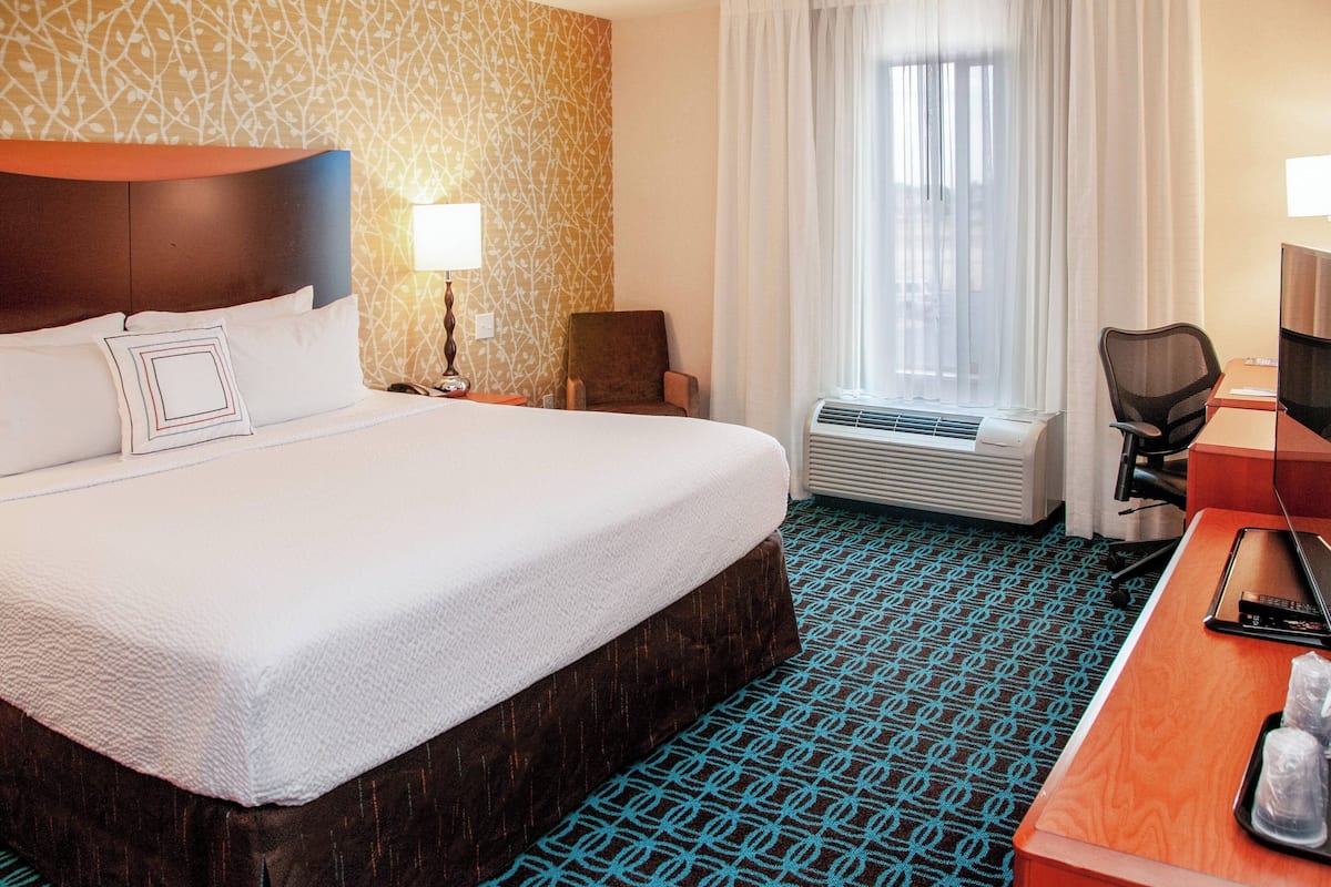 A hotel room with a large bed, a desk, a chair, a lamp, an air conditioning unit, and a window with curtains.