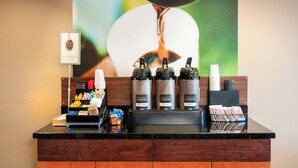 A coffee station with three coffee dispensers labeled "Buzz Worthy" and "Hot Water," a coffee cup holder, and a tray with various coffee pods and a small basket of pastries. - Fairfield Inn & Suites Colorado Springs N./Air Force Academy (Colorado Springs)