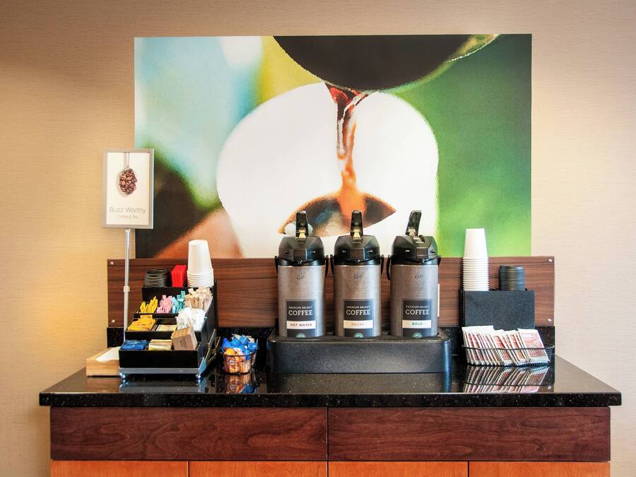 Un espace dédié au café, comprenant trois distributeurs de café étiquetés « Café à ne pas manquer » et « Eau chaude », un porte-tasse et un plateau avec divers dosettes de café et un petit panier de viennoiseries.