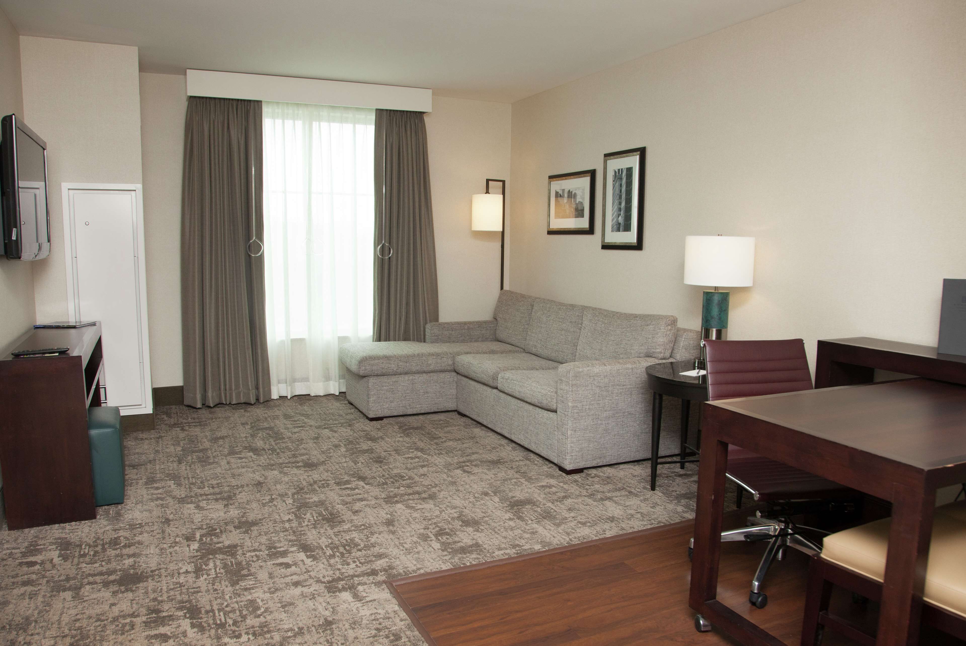 Habitación de hotel con sofá modular, una mesita con lámpara, escritorio con silla, televisor montado en la pared y una puerta.