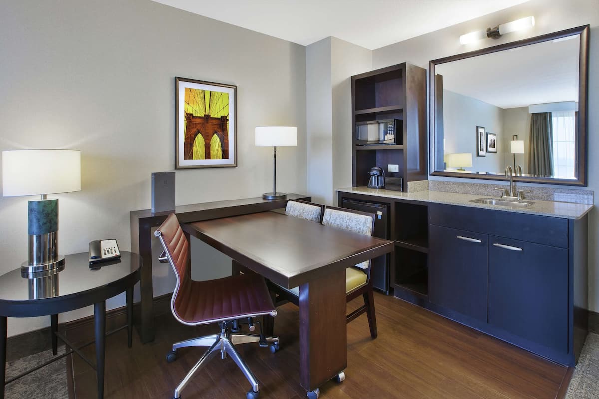 Quarto de hotel com uma mesa de conferência, uma cadeira, escrivaninha com luminária, micro-ondas, cafeteira, pia e um espelho grande.