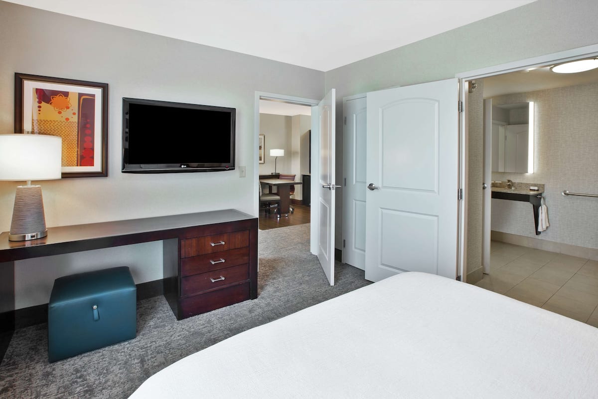 Quarto de hotel com cama, escrivaninha com luminária, televisão e banheiro com espelho.