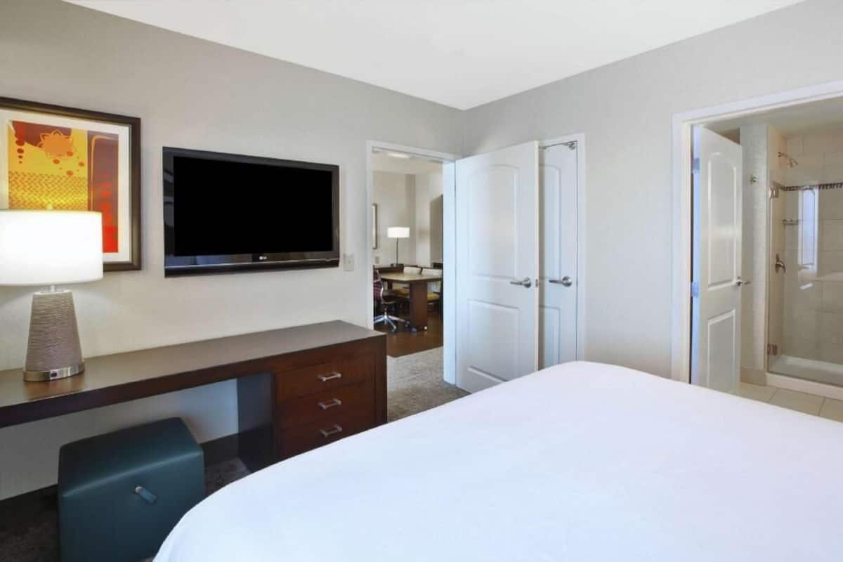 Quarto de hotel com cama, televisão, escrivaninha com gavetas, luminária, mala e área de chuveiro.