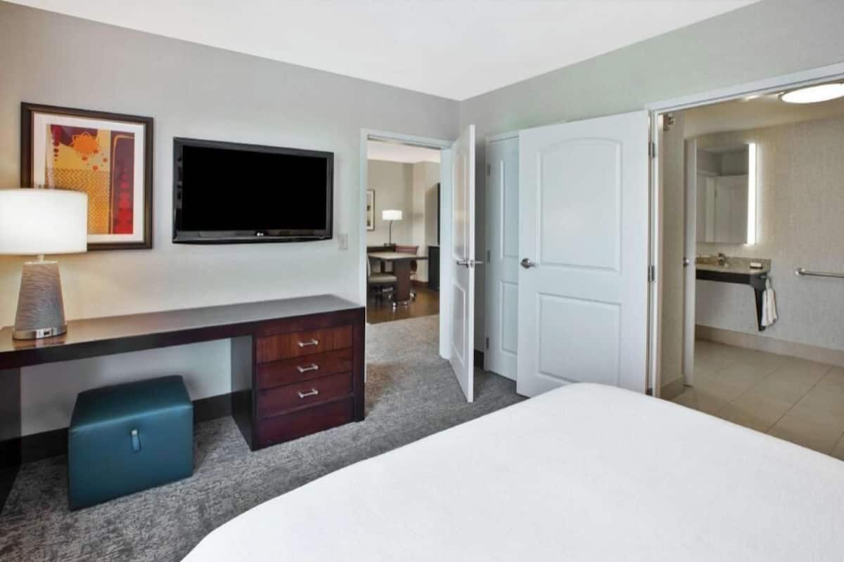 Quarto de hotel com cama, escrivaninha com luminária, televisão, guarda-roupa e banheiro visível através de uma porta aberta.