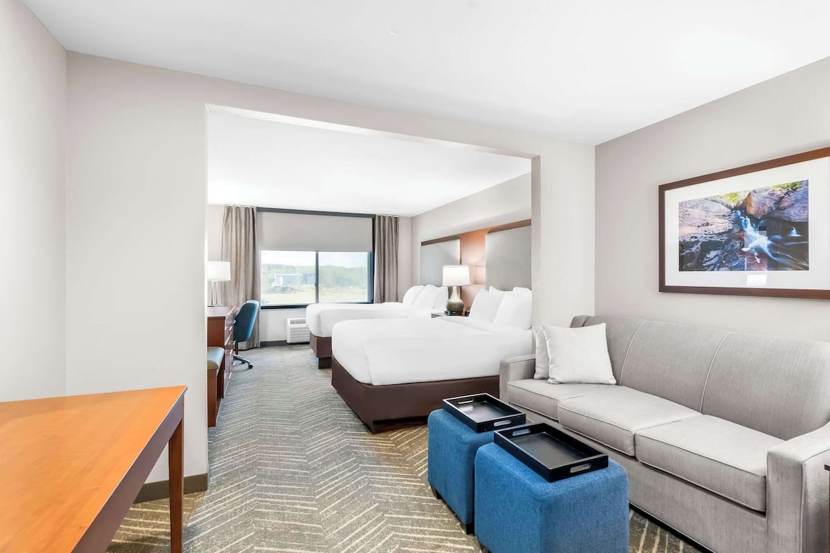 A hotel room with a large bed, a sofa, a desk, and a chair. There is a window with curtains, a lamp, and a framed picture on the wall.