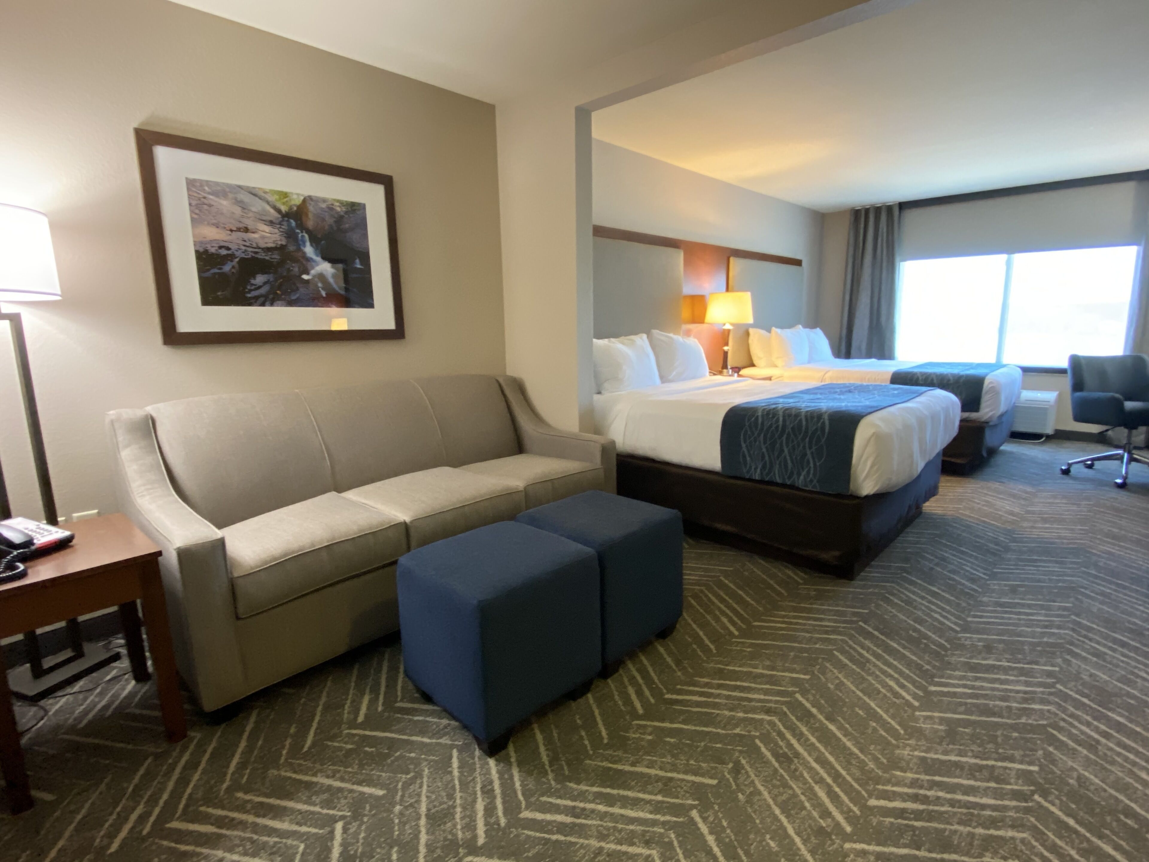 A hotel room with a sofa, two ottomans, a bed with a blue blanket, a desk with a telephone, and a framed picture on the wall.