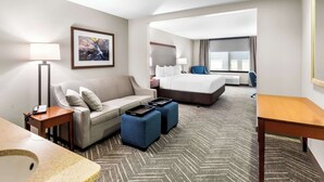 A hotel room with a sofa, two ottomans, a bed, a desk, a lamp, a framed picture, and a window.