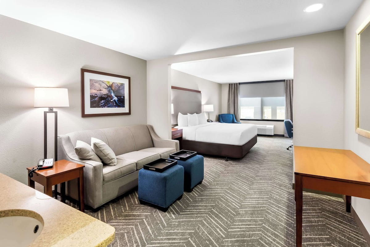 A hotel room with a sofa, two ottomans, a bed, a desk, a lamp, a framed picture, and a window.