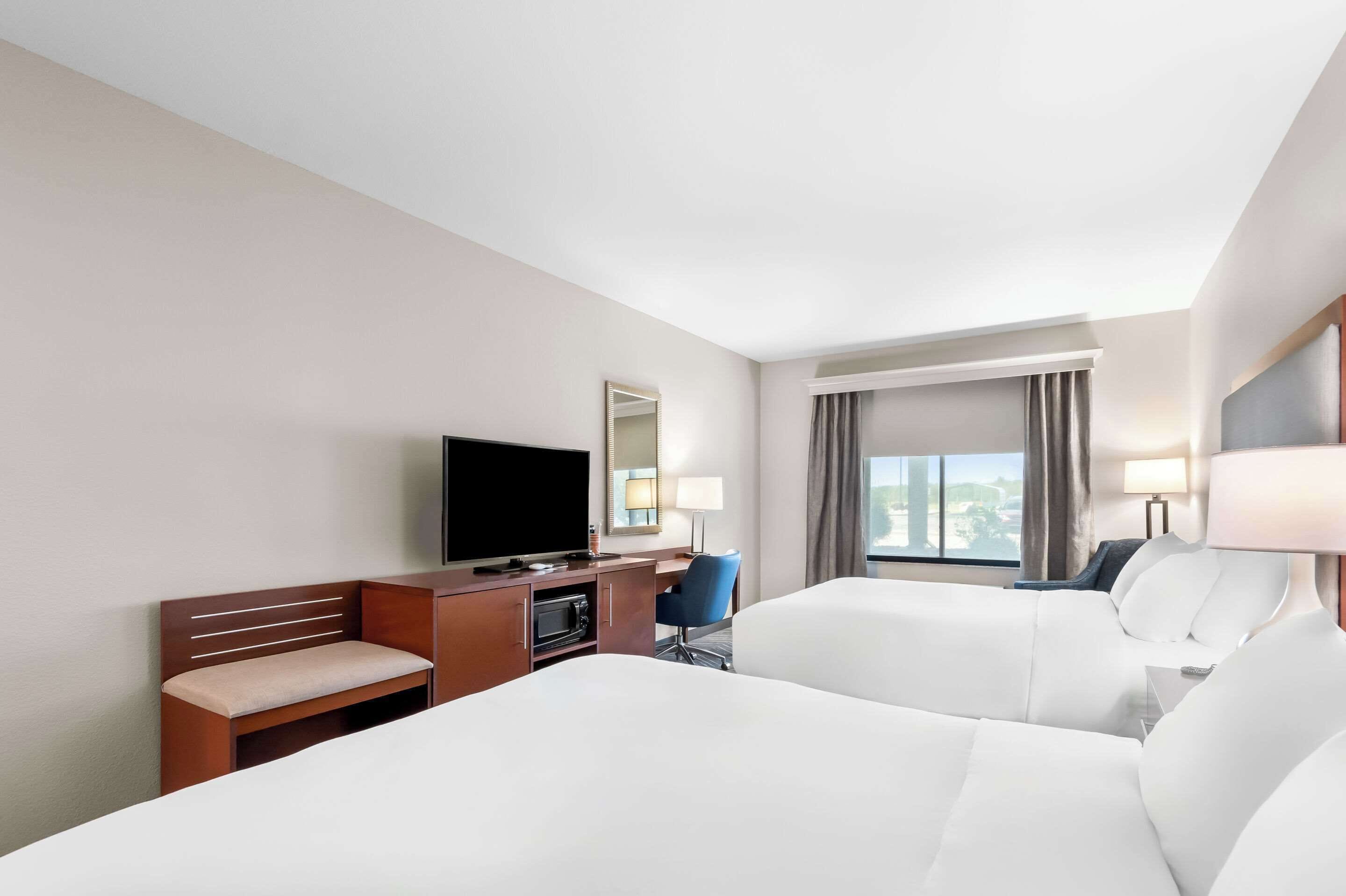 Ein Hotelzimmer mit zwei Betten, einem Fernseher, einem Schreibtisch mit Stuhl und einem fensterbepflanzten Fenster.