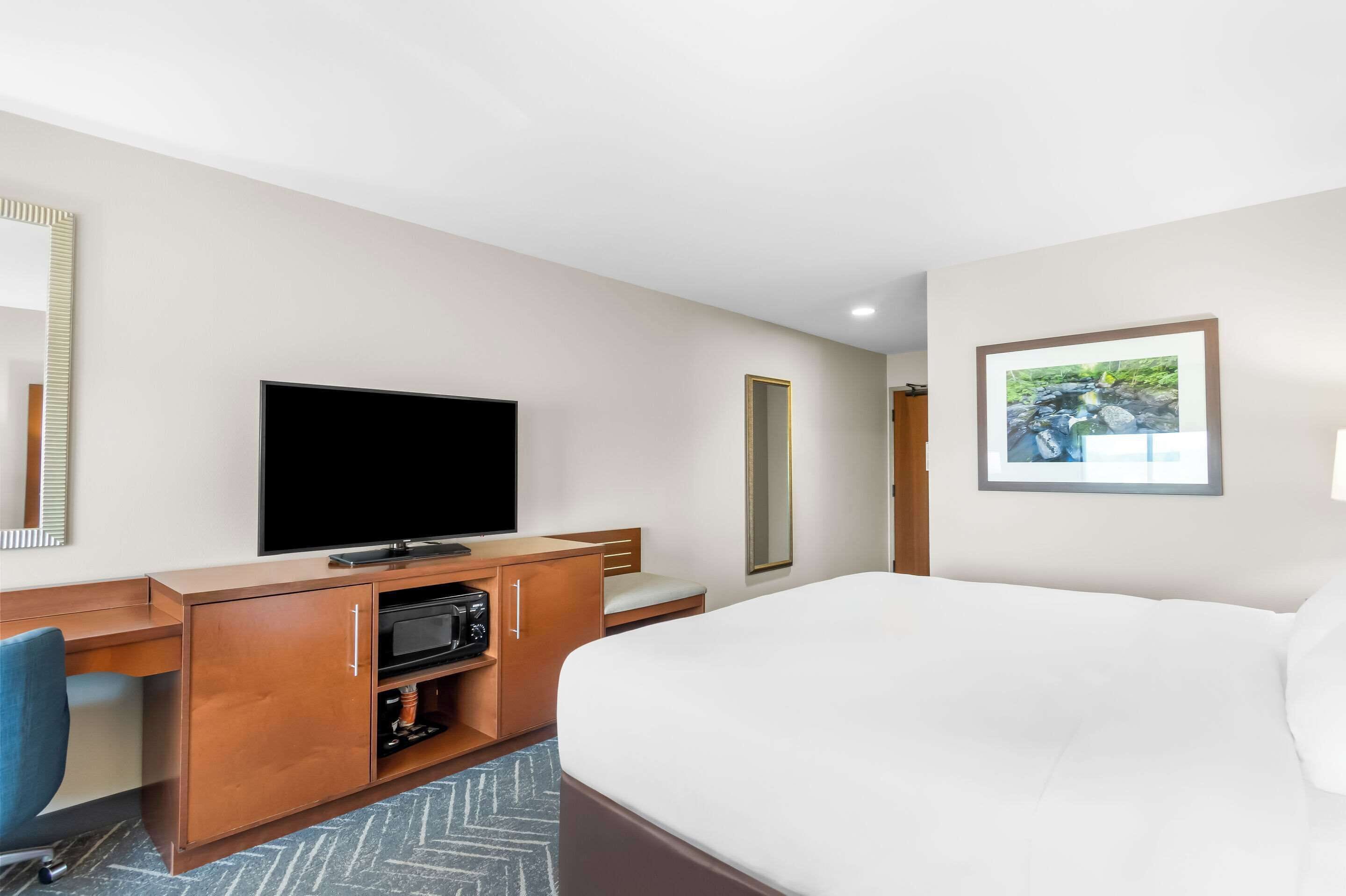 Ein Hotelzimmer mit einem großen Bett, einem Fernseher, einem Schreibtisch mit Stuhl, einem Spiegel und einem gerahmten Bild an der Wand.
