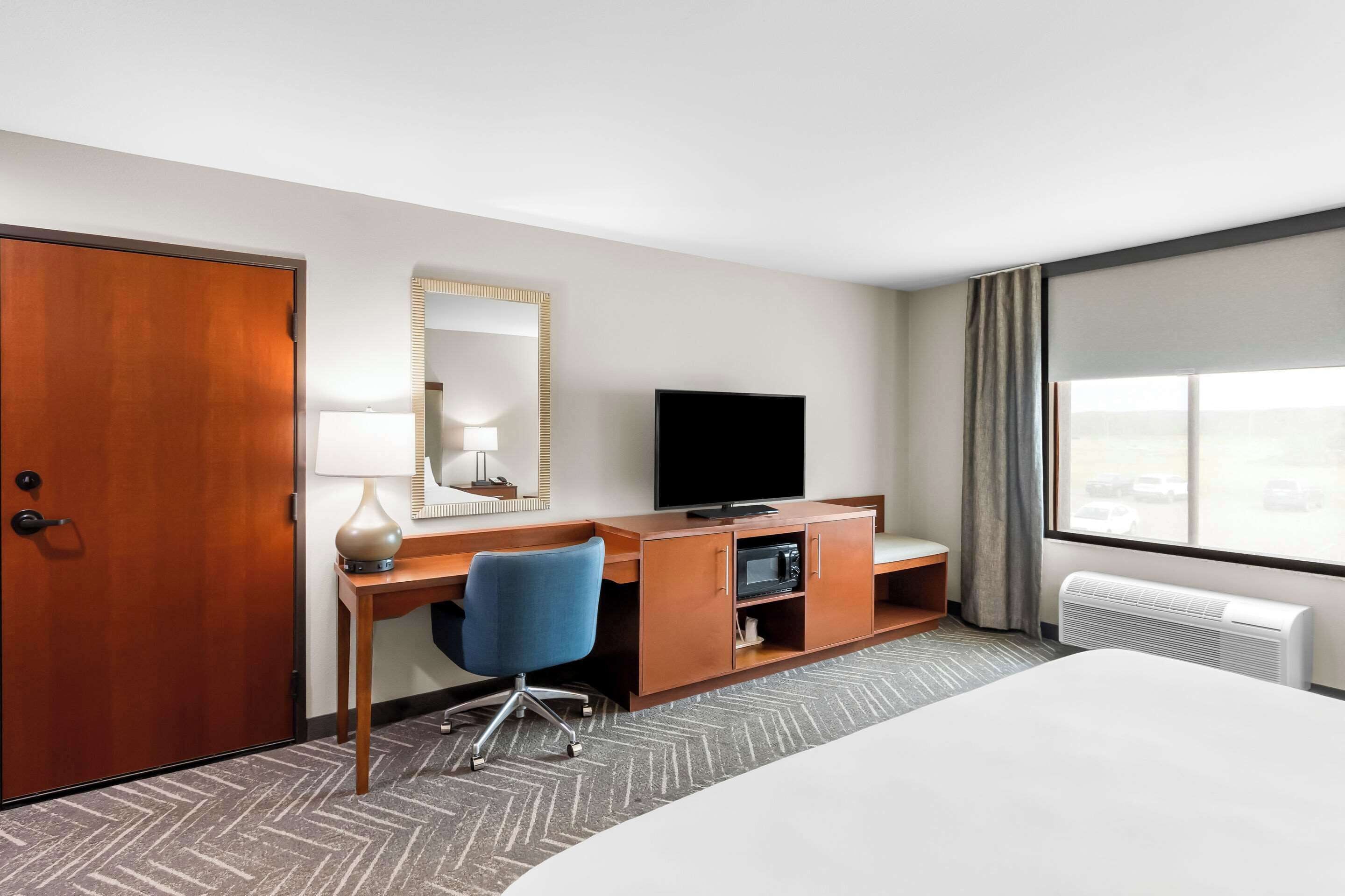 Ein Hotelzimmer mit einem Bett, einem Schreibtisch mit Computer, einem Fernseher, einem Spiegel und einem Fenster mit Vorhängen.