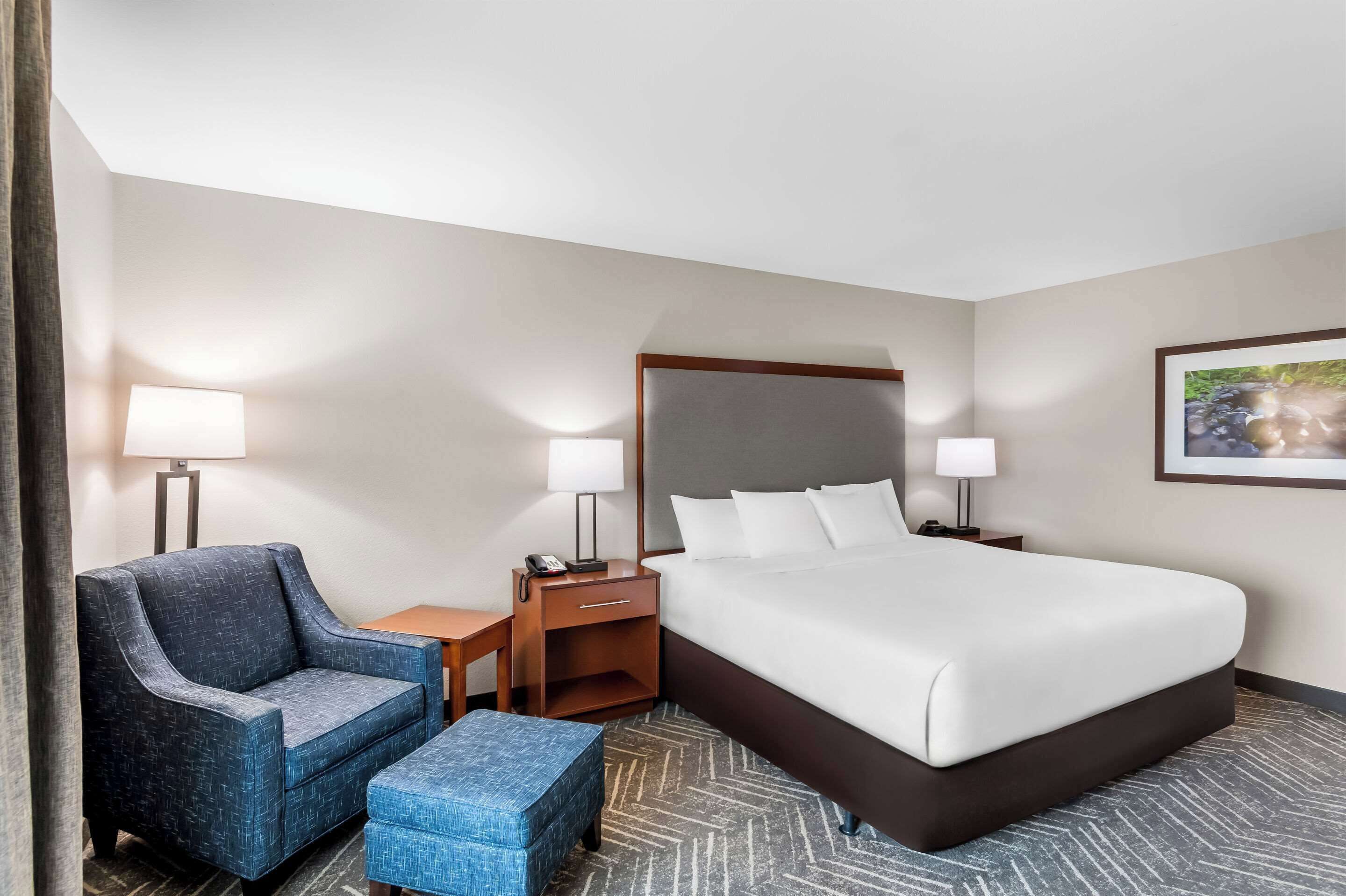 A hotel room with a bed, a chair, a desk, a lamp, and a nightstand.
