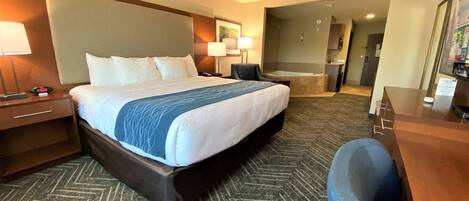 A hotel room with a large bed, a desk, a chair, a desk lamp, a nightstand, a wall-mounted TV, a bathroom with a bathtub, and a sink.