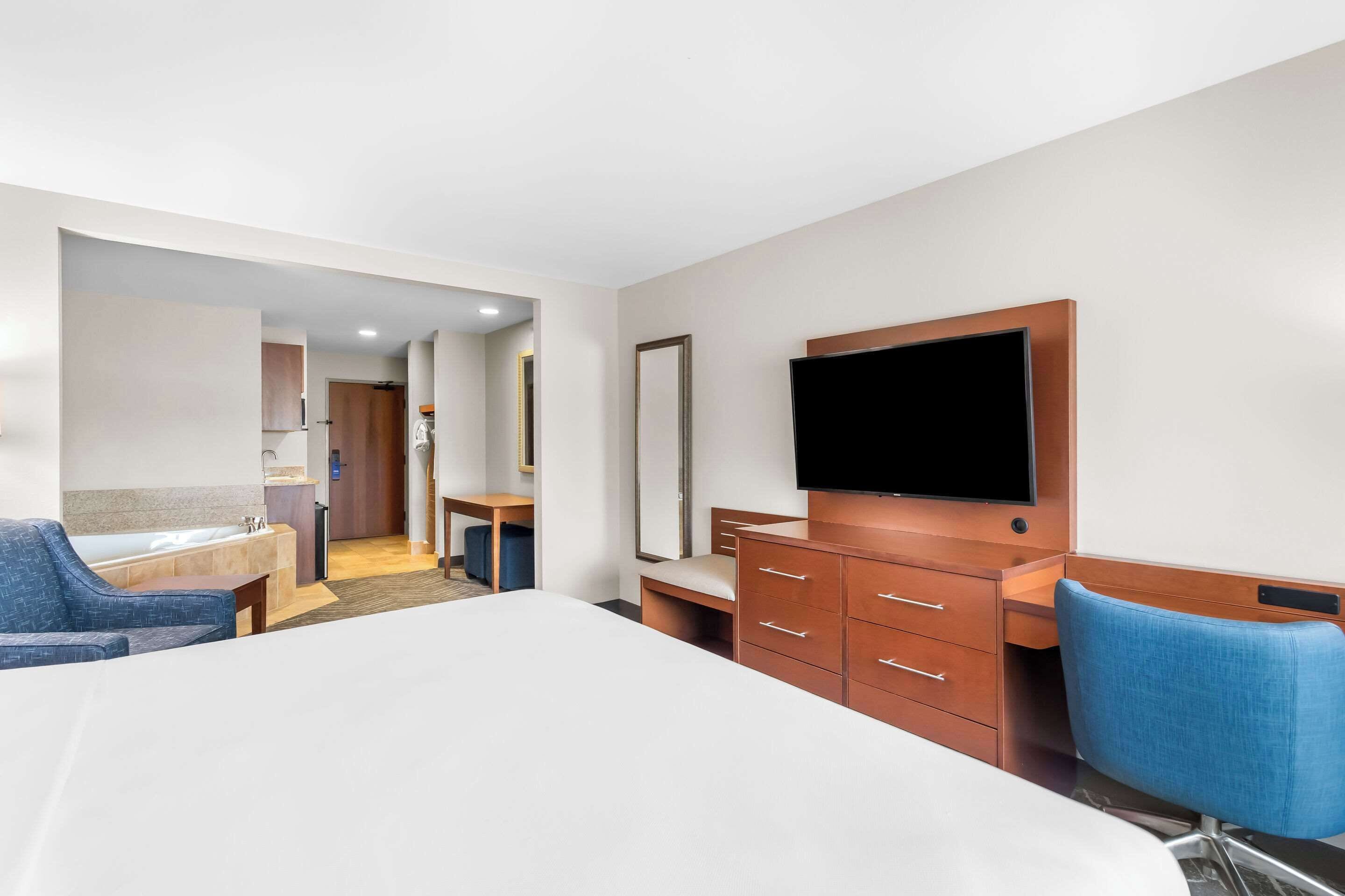 A hotel room with a bed, a desk, a chair, a TV, a dresser, and a bathroom visible through an open door.