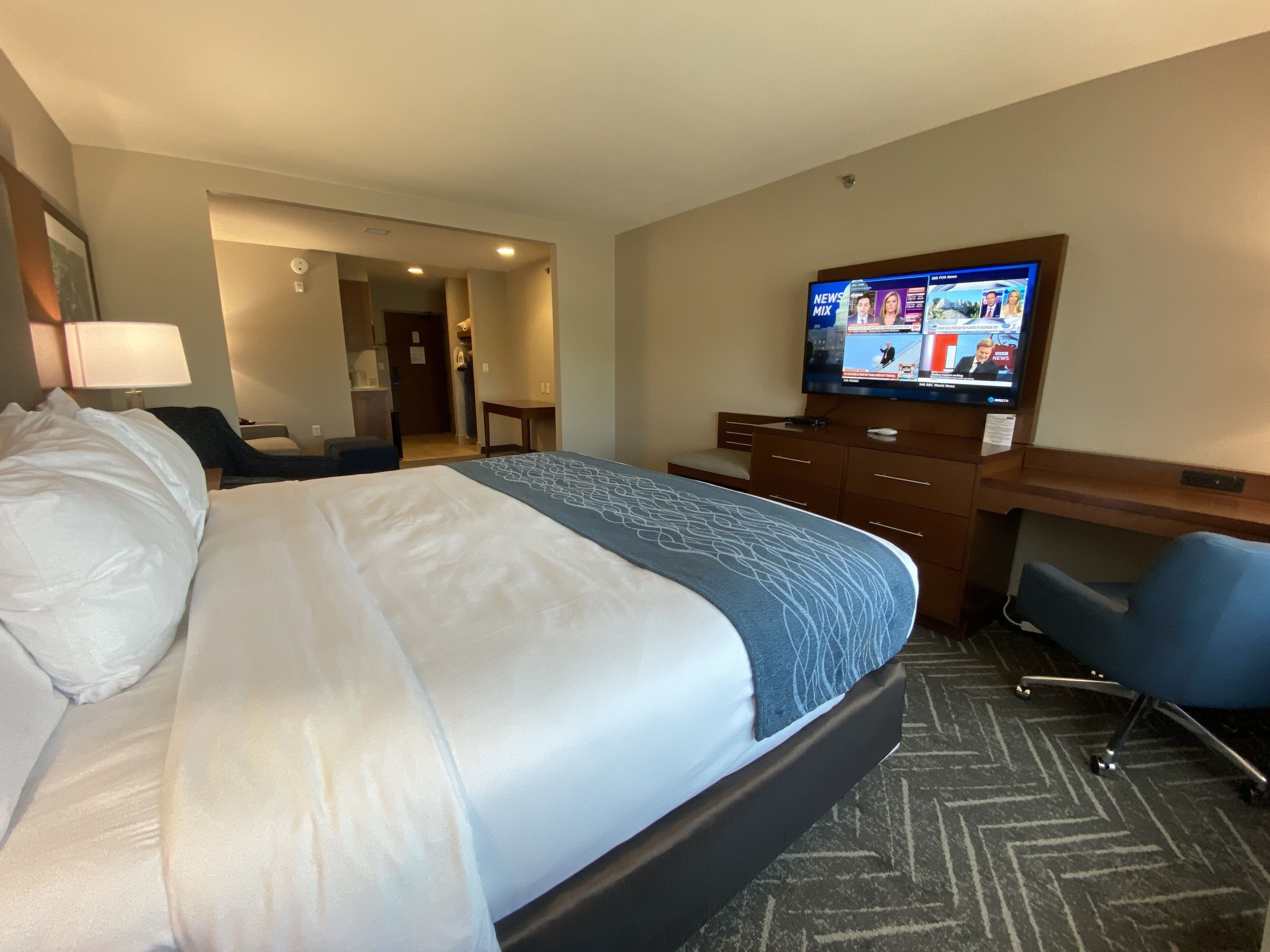 A hotel room with a large bed, a desk, a chair, and a television displaying news.
