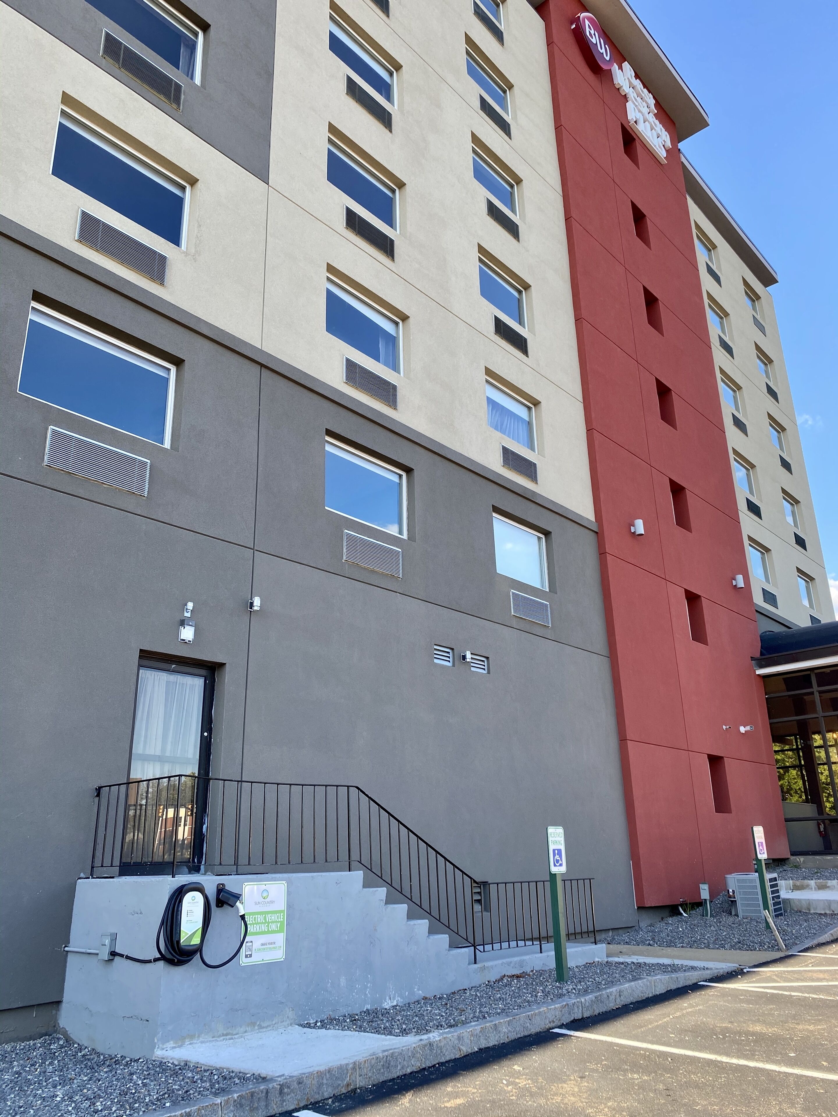 Um hotel de vários andares com fachada em vermelho e cinza, grandes janelas e área de estacionamento com estações de carregamento para veículos elétricos.