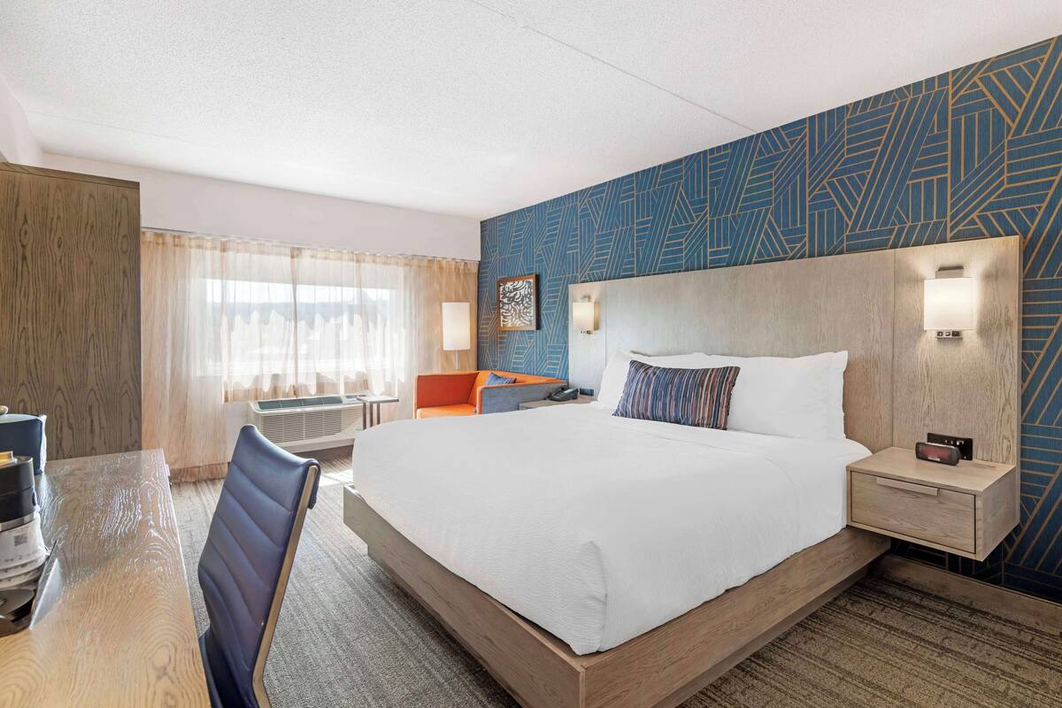 A hotel room with a large bed, a desk, and a chair. There is a window with curtains, a wall with a geometric pattern, and a nightstand with a lamp.
