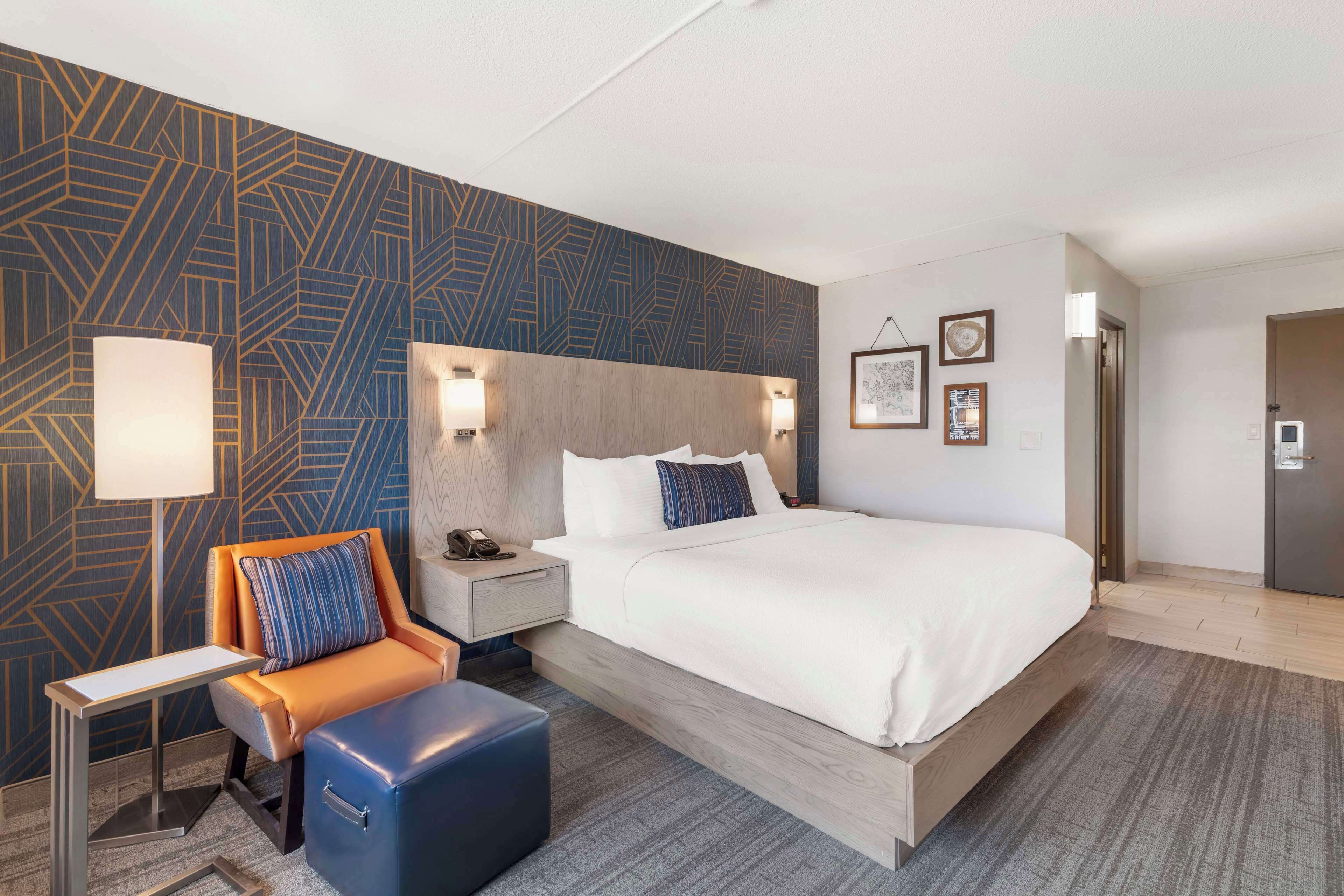 Ein Hotelzimmer mit einem großen Bett, einem Nachttisch mit Lampe, einem orangefarbenen Sessel, einem blauen Hocker und einer Wand mit geometrischem Muster.