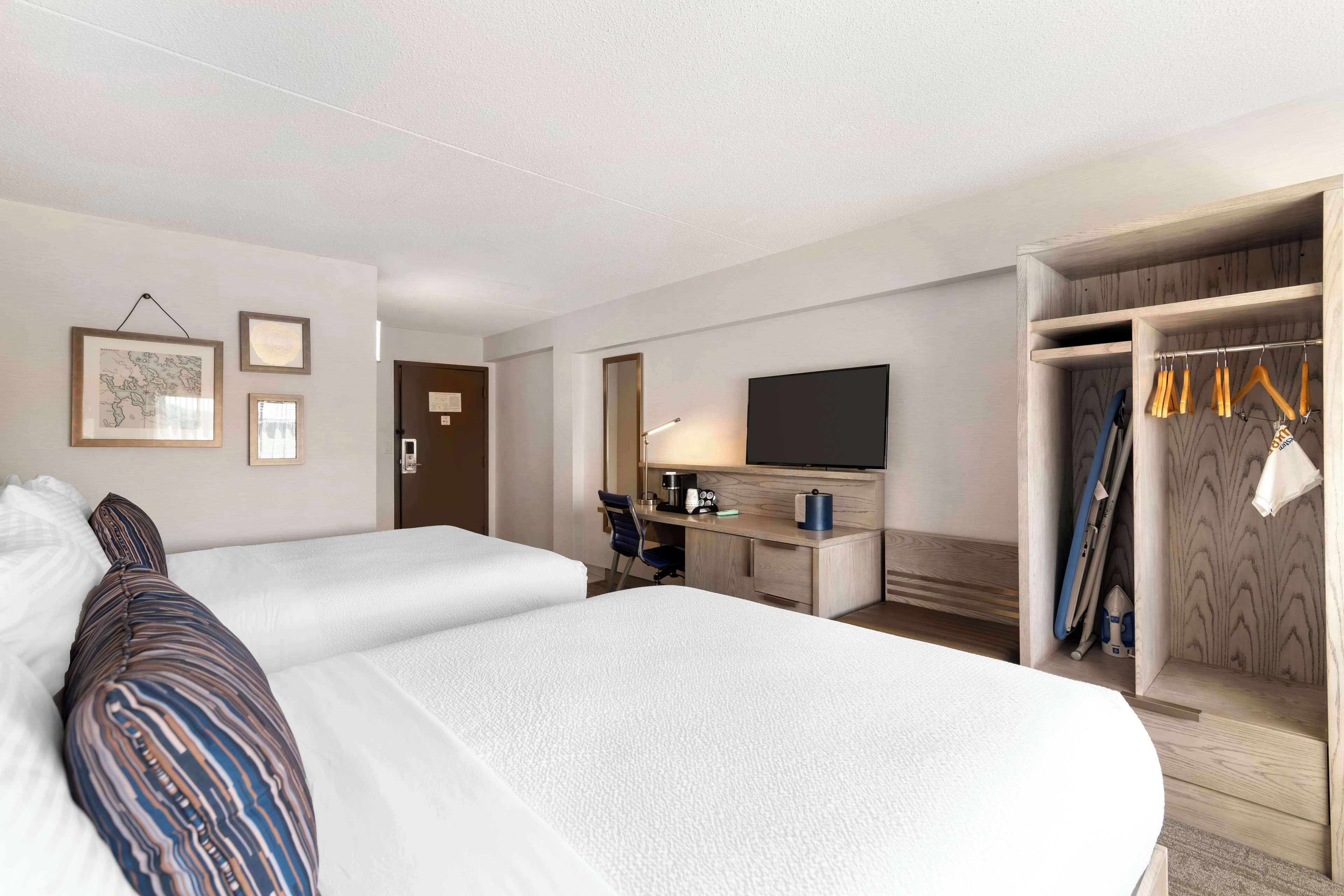 Quarto de hotel com duas camas, uma mesa de trabalho com televisão, guarda-roupa e cabideiro.