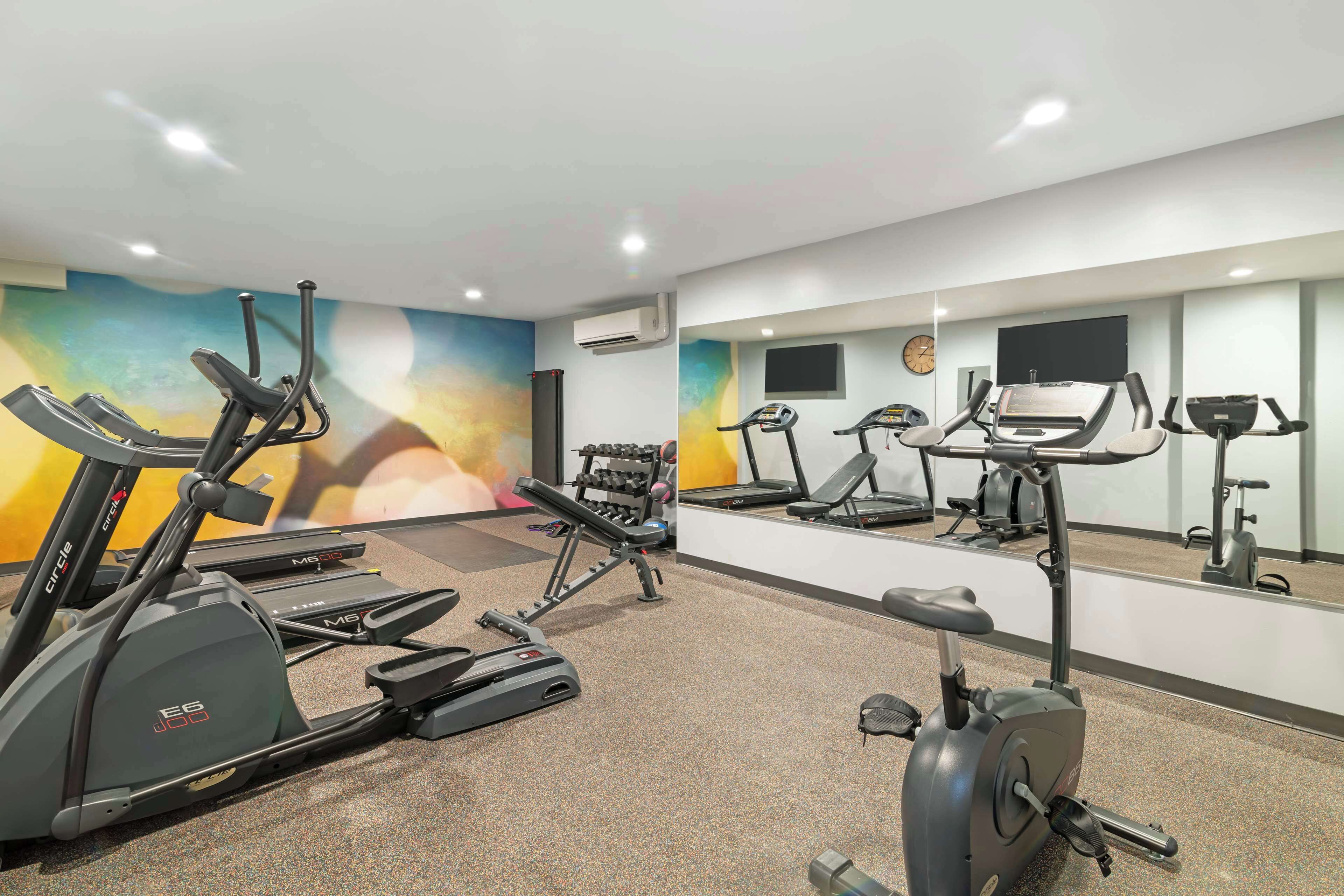 Une salle de sport équipée de plusieurs appareils d’exercice, d’un grand miroir et d’une horloge au mur.