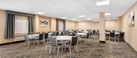 Un salón de conferencias con varias mesas y sillas, piso alfombrado y ventanas grandes con cortinas.