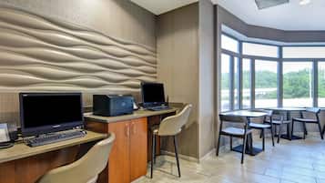 Un área de recepción de hotel moderna con dos estaciones de trabajo de computadora, un teléfono e impresora. Hay varias sillas y una mesita. La habitación tiene ventanas amplias que ofrecen vistas al verde exterior.