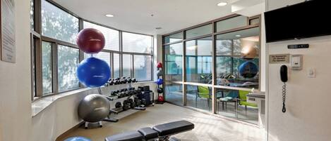 Un gimnasio con equipamiento de ejercicio, una banca y ventanas amplias.