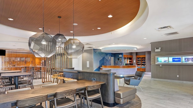 Un hall d’hôtel moderne avec un comptoir d’accueil en bois, des luminaires suspendus hexagonaux et un espace salon avec des tables et des chaises.